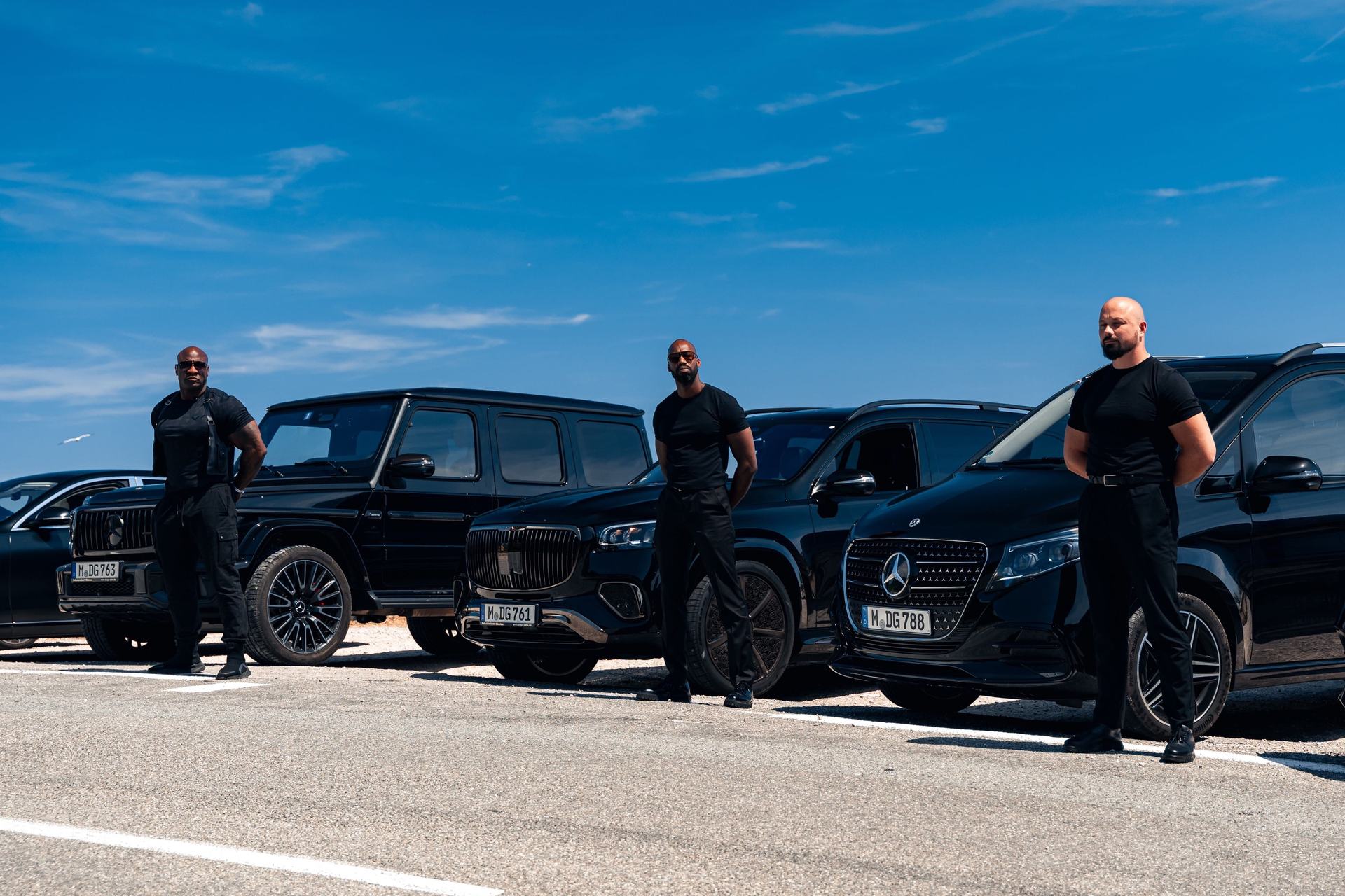 Three LEON security drivers standing in front of black Mercedes vehicles under a blue sky